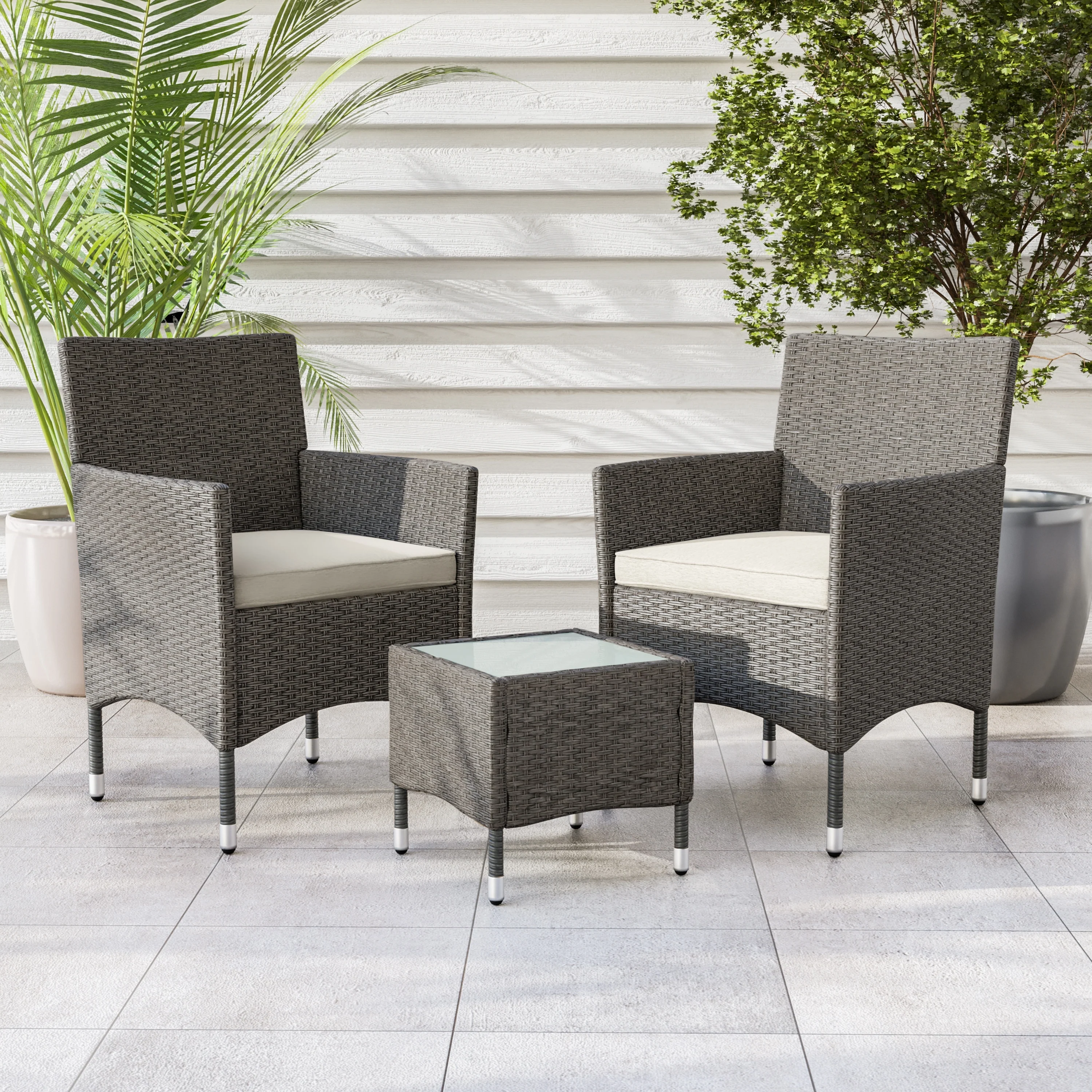Outdoor patio set with two gray wicker chairs and a square glass-top side table, all featuring cream cushions. Lush green plants in large pots are placed in the background.