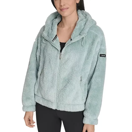 A woman wearing a light blue, fluffy hooded jacket from Calvin Klein with zippered pockets, paired with black leggings.