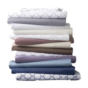 A stack of Liz Claiborne sheet sets in various colors and patterns, including white, gray, beige, pink, brown, purple, and blue, with some featuring a decorative interlocking design.