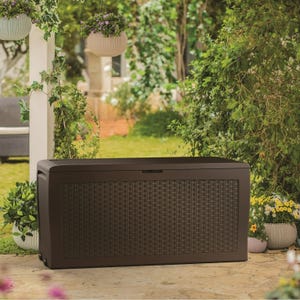 A brown, wicker-patterned, 80-gallon deck box is set in a garden, offering outdoor storage and seating solutions.