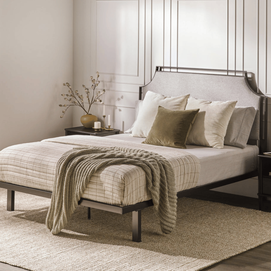 A bedroom features a metal bed frame with a neutral-toned bedspread, accentuated by a textured throw and green pillows. A nightstand holds a decorative vase with branches and candles, set against a paneled wall.