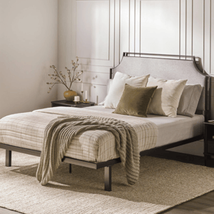 A bedroom features a metal bed frame with a neutral-toned bedspread, accentuated by a textured throw and green pillows. A nightstand holds a decorative vase with branches and candles, set against a paneled wall.