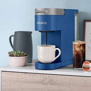 A blue Keurig coffee maker brewing into a white mug, beside a grey pitcher, a small potted plant, and an iced coffee in a glass.