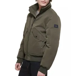 A man wears an olive green bomber jacket with a zip front, two large flap pockets, and a patch on the left sleeve, paired with dark jeans.