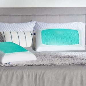 Two pillows with green cooling gel pads are displayed on a bed with a gray headboard and gray bedding, accompanied by patterned decorative pillows.