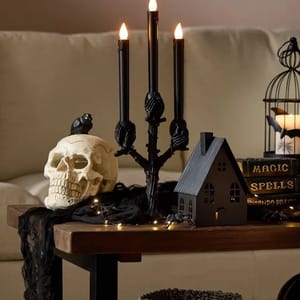 A skull, a candelabra with lit candles, a small house decoration, a book titled \“Magic Spells,\“ and string lights on a table.