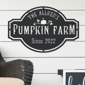A decorative sign for \“The Allred's Pumpkin Farm\“ features a pumpkin and floral design, with the text \“Since 2022\“ on a horizontal wooden panel background, alongside a wicker chair and part of another framed item.