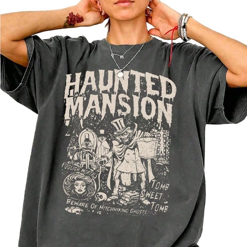 A person is wearing an oversized black T-shirt featuring a \“Haunted Mansion\“ graphic with spooky illustrations and phrases like \“Tomb Sweet Tomb\“ and \“Beware of Hitchhiking Ghosts.\“ They accessorize with necklaces and bracelets.