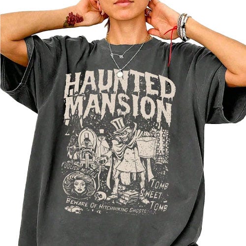 A person is wearing an oversized black T-shirt featuring a \“Haunted Mansion\“ graphic with spooky illustrations and phrases like \“Tomb Sweet Tomb\“ and \“Beware of Hitchhiking Ghosts.\“ They accessorize with necklaces and bracelets.