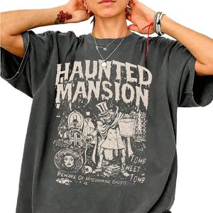 A person is wearing an oversized black T-shirt featuring a \“Haunted Mansion\“ graphic with spooky illustrations and phrases like \“Tomb Sweet Tomb\“ and \“Beware of Hitchhiking Ghosts.\“ They accessorize with necklaces and bracelets.