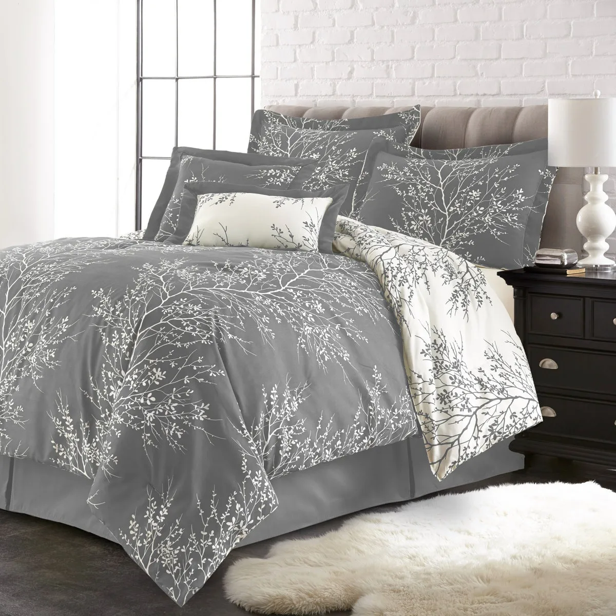 A stylish bedding set with a gray duvet cover and matching pillows, featuring a white branch and leaf pattern, is displayed on a bed with a tufted headboard. A black nightstand and white table lamp are beside the bed, and a fluffy white rug is on the floor.