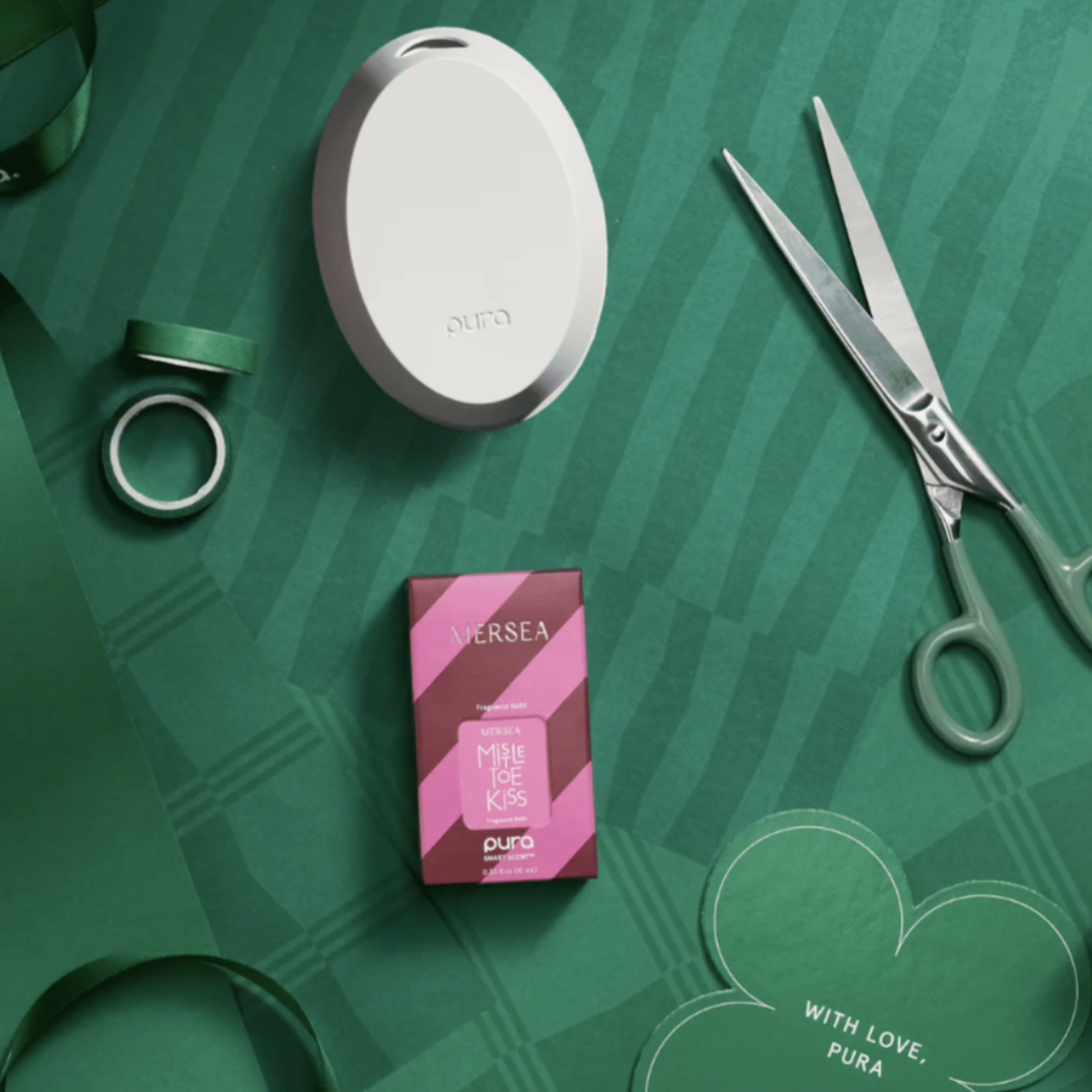 A Pura home fragrance diffuser and scent vial labeled \“Mistletoe Kiss\“ by Mersea are placed on a green, festive background with ribbon, wrapping paper, and scissors.
