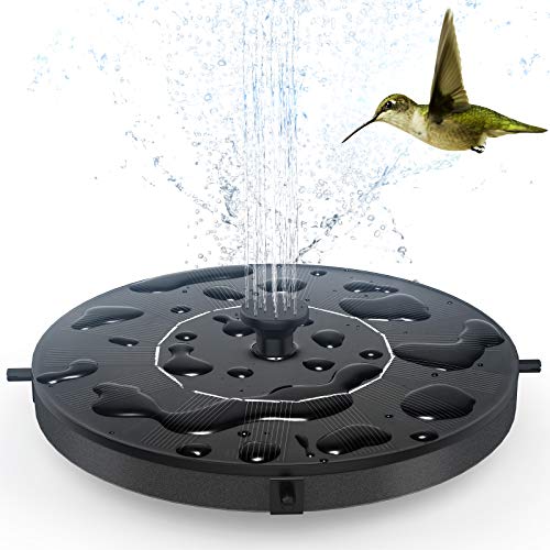 A circular, black solar-powered fountain with multiple spouts is spraying water upwards, and a hummingbird is flying nearby.
