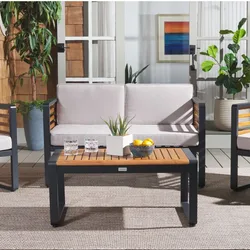 Outdoor patio set featuring a couch with light cushions, a wooden coffee table with black metal frame, two glasses, a small plant, a bowl of lemons, and potted plants in the background.