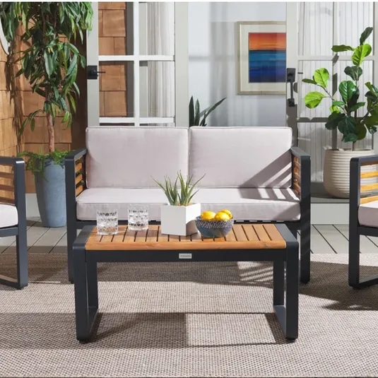 Outdoor patio set featuring a couch with light cushions, a wooden coffee table with black metal frame, two glasses, a small plant, a bowl of lemons, and potted plants in the background.
