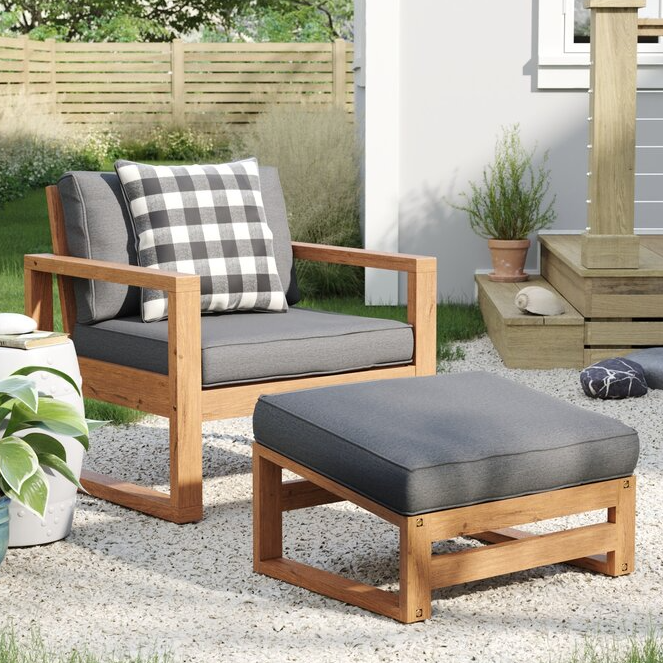 A wooden outdoor armchair with gray cushions and a matching ottoman is shown. The chair features a black and white checkered pillow, set on a pebble-filled patio next to a small plant.