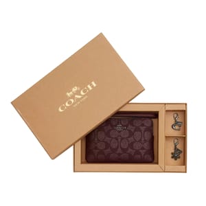 Coach Boxed Wristlet includes a monogram-patterned wristlet in maroon and two charm keychains, one shaped like a letter 'C' with a clip and the other a snowflake. Packaged in a beige box with the Coach logo on the lid.