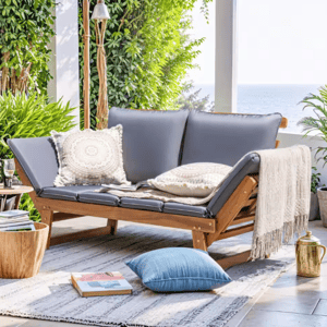 A wooden outdoor sofa with adjustable backrests and gray cushions, adorned with patterned pillows and a beige throw, is set in a lush, sunlit patio with surrounding plants and decor, including a rug, side table, and a blue pillow on the floor.