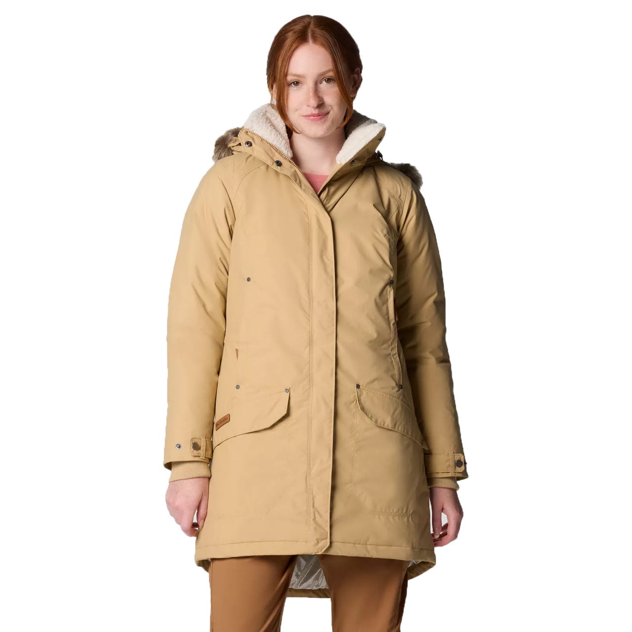 A woman is wearing a tan winter coat with a hood lined in faux fur, featuring front pockets and button details. The coat is paired with matching tan pants.