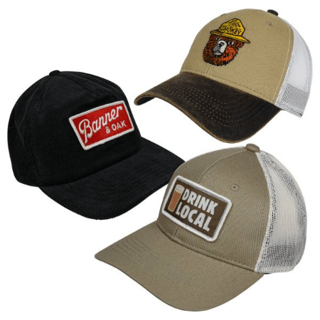 Three trucker hats are shown: a black one with \“Banner & Oak\“ in red, a khaki one with \“Drink Local\“ and a beer glass, and another khaki hat featuring an embroidered bear wearing a hat labeled \“Smokey,\“ with a brown distressed bill.