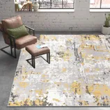 Gray and yellow abstract-patterned rug in a modern room with a brown chair, green cushion, ottoman, and a potted cactus on a concrete floor.