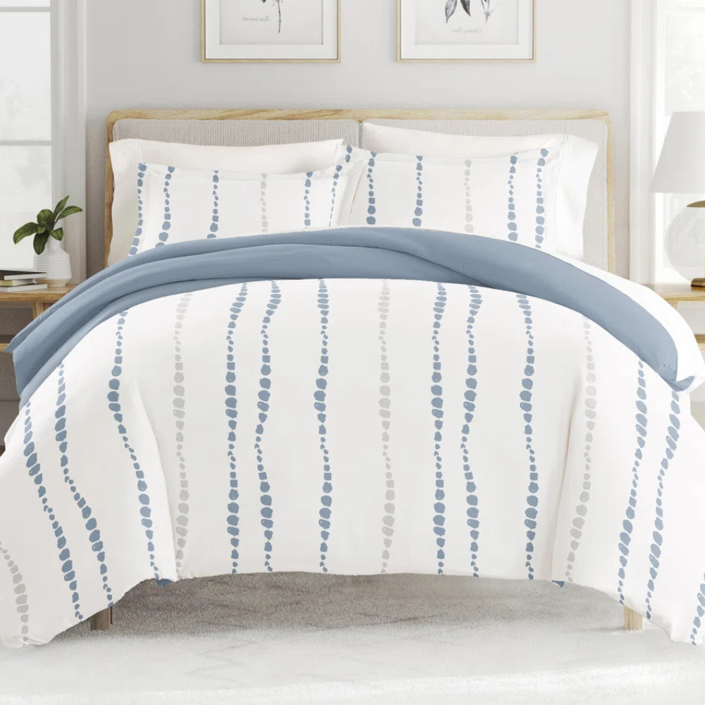 A bed is shown with a blue and white patterned duvet cover featuring vertical dotted lines. The bedding set includes matching pillowcases, and the room is styled with simple, neutral decor.