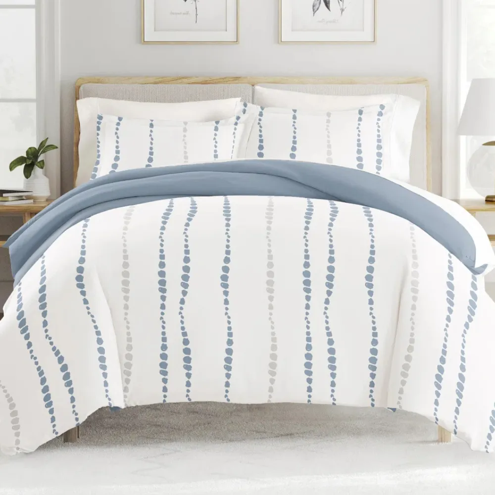 A bed is shown with a blue and white patterned duvet cover featuring vertical dotted lines. The bedding set includes matching pillowcases, and the room is styled with simple, neutral decor.