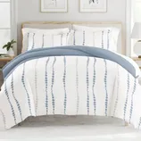 A bed is shown with a blue and white patterned duvet cover featuring vertical dotted lines. The bedding set includes matching pillowcases, and the room is styled with simple, neutral decor.