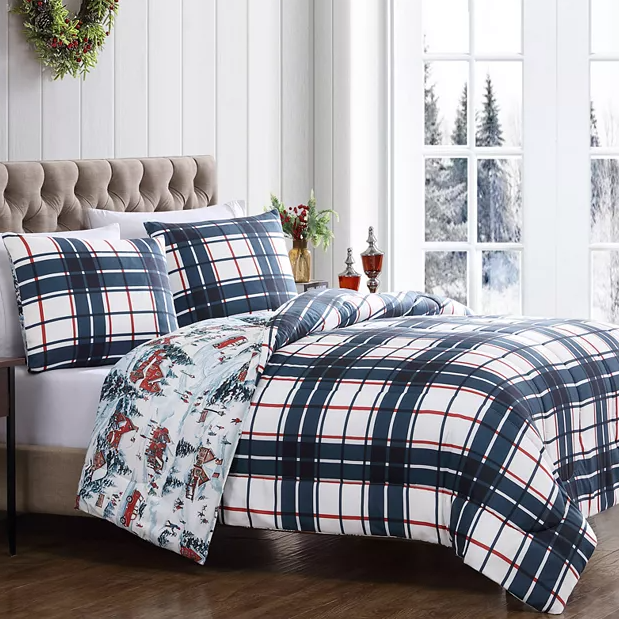A bed features a plaid-patterned duvet and pillow set, complemented by a reversible design with a winter village motif. The room has a cozy, festive atmosphere with a wreath and snow-covered window background.