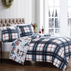 A bed features a plaid-patterned duvet and pillow set, complemented by a reversible design with a winter village motif. The room has a cozy, festive atmosphere with a wreath and snow-covered window background.