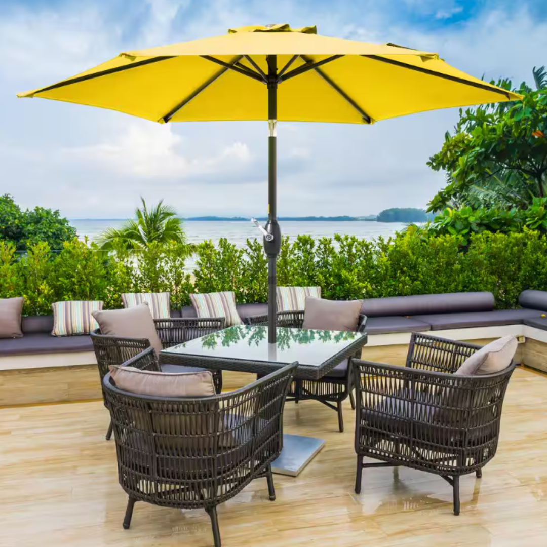 A 7.5' yellow patio market umbrella with a central pole is set over a table surrounded by wicker chairs, placed on a wooden deck with a view of greenery and water in the background.