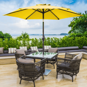 A 7.5' yellow patio market umbrella with a central pole is set over a table surrounded by wicker chairs, placed on a wooden deck with a view of greenery and water in the background.