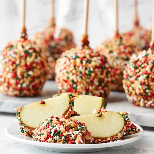 Twelve caramel apples coated in multicolored sprinkles, presented with wooden sticks as handles, with three apple slices dipped in the same coating in the foreground.