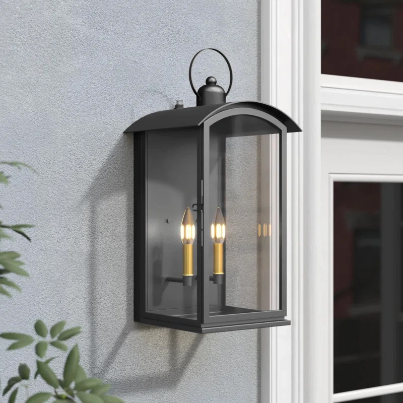 The Lark Manor Lantern is a wall-mounted outdoor light fixture with a black metal frame and glass panels, featuring two candle-style bulbs inside. It has a classic lantern design with an arched top and handle.