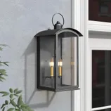 The Lark Manor Lantern is a wall-mounted outdoor light fixture with a black metal frame and glass panels, featuring two candle-style bulbs inside. It has a classic lantern design with an arched top and handle.