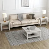 Living room setup with a beige sofa, two side tables with lamps, a central coffee table holding a laptop, phone, and cup, and a patterned rug on wooden flooring.