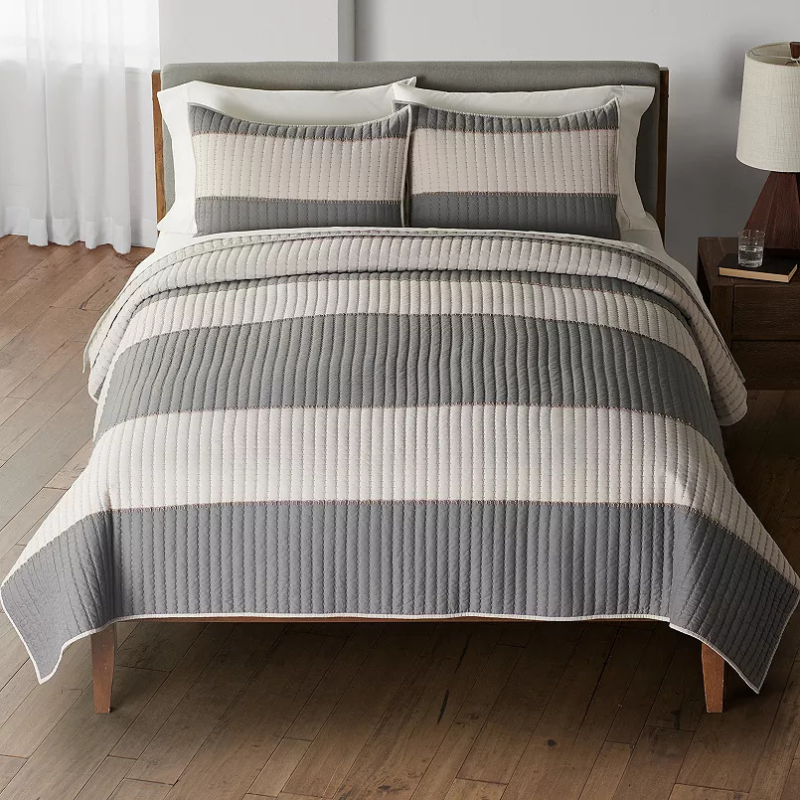 A bed with a gray and white striped quilt and matching pillow shams, situated on a wooden floor with a bedside table and lamp.