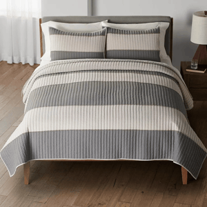 A bed with a gray and white striped quilt and matching pillow shams, situated on a wooden floor with a bedside table and lamp.