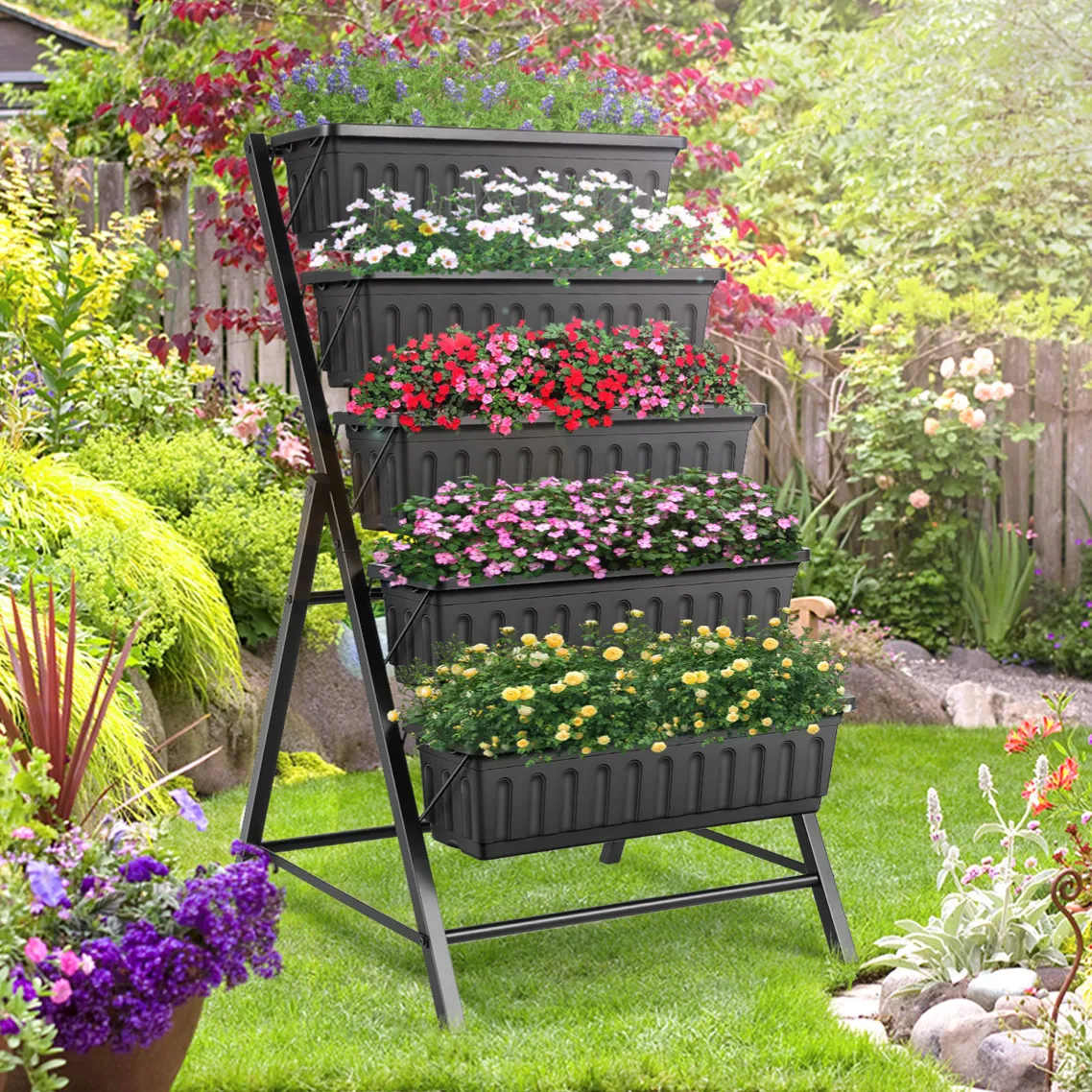 A multi-tier vertical garden planter with five levels, each filled with colorful flowers, is displayed in a lush garden setting with various plants and a wooden fence in the background.
