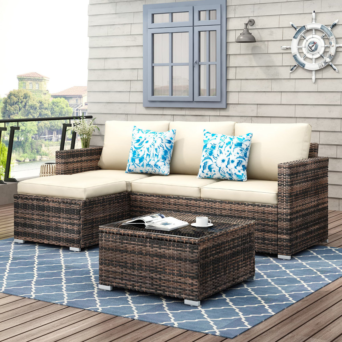 All-weather sectional sofa set with two-tone wicker and cream cushions, featuring blue floral accent pillows. Includes a matching wicker coffee table with a glass top on a blue patterned outdoor rug, set on a wooden deck with beach-themed decor.