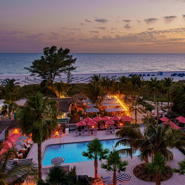 Aerial view of a beachside resort with a swimming pool, umbrellas, palm trees, and a lit-up dining area during twilight.