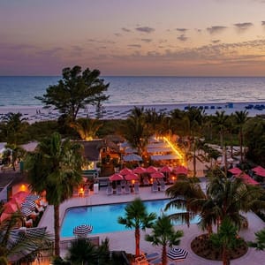 Aerial view of a beachside resort with a swimming pool, umbrellas, palm trees, and a lit-up dining area during twilight.