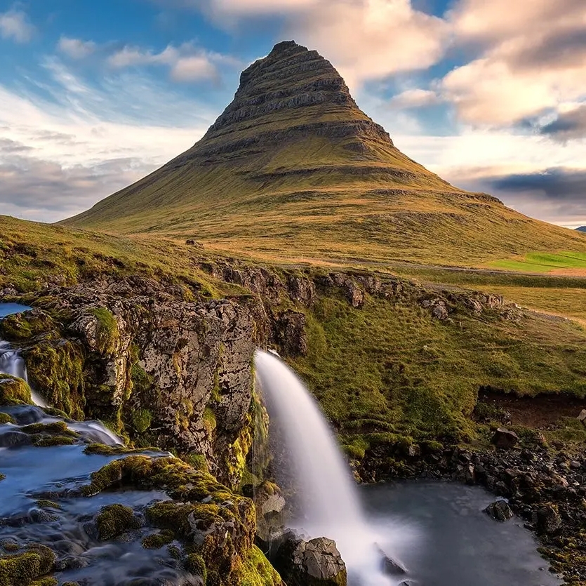 A picturesque landscape featuring Kirkjufell mountain with a cascade waterfall in the foreground, under a partly cloudy sky.