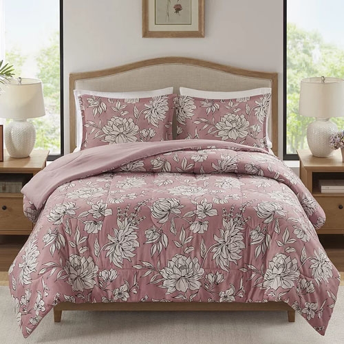 A bed features a floral-patterned quilt in pink with matching pillow shams. The bedding is accented by a light beige headboard and is surrounded by wooden nightstands with table lamps.