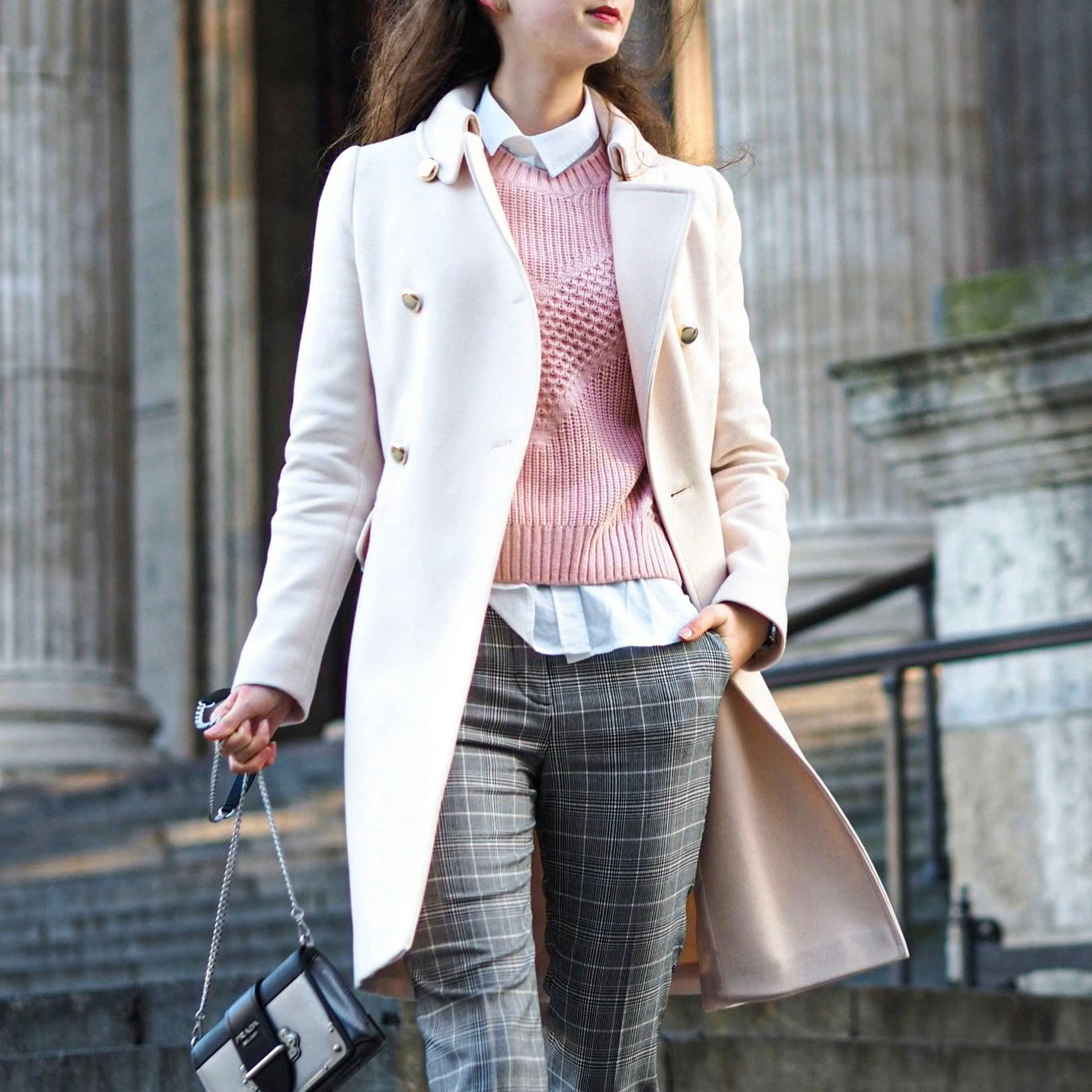 A person wearing a white coat, pink sweater, checkered trousers, with a white shirt collar visible and carrying a chain-strap black bag and a large taupe tote.