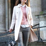 A person wearing a white coat, pink sweater, checkered trousers, with a white shirt collar visible and carrying a chain-strap black bag and a large taupe tote.