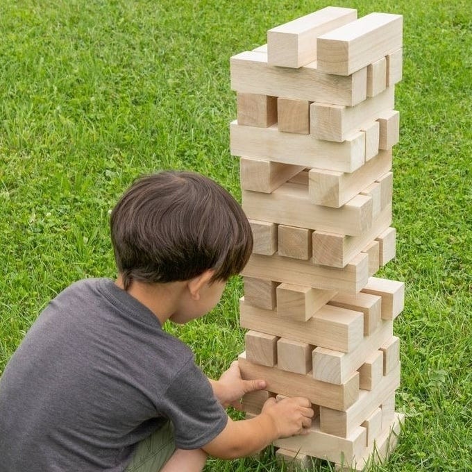 A child is playing a game of giant Jenga on a grass lawn, removing a block from a tall stacked tower of large wooden pieces.
