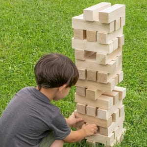 A child is playing a game of giant Jenga on a grass lawn, removing a block from a tall stacked tower of large wooden pieces.