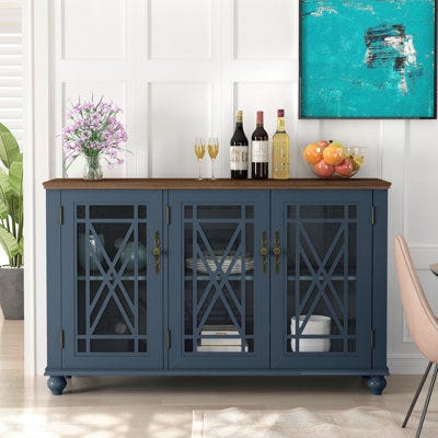 A blue three-door wooden cabinet with a brown top, displaying bottles, glasses, and a bowl of fruit, against a white paneled wall with a blue abstract painting above.