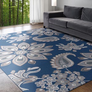 A blue area rug with large floral and paisley designs is placed in a modern living room with a gray sofa, a floor lamp, and a floor-to-ceiling window looking out to a green forest.